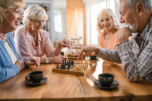 Elderly couples smiling and playing chess at a wooden table during afternoon tea.