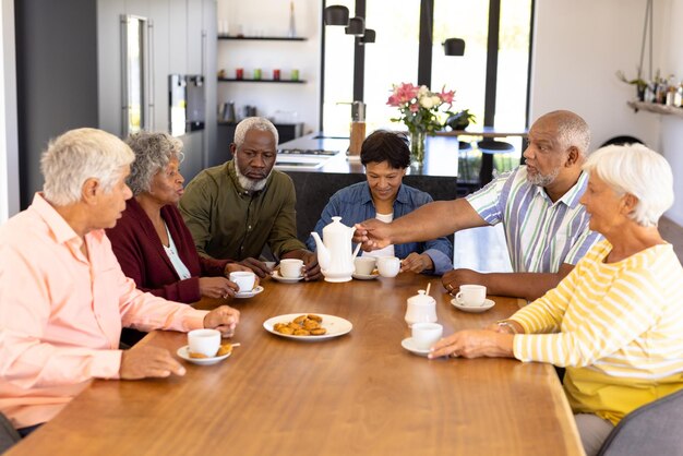 A group of senior friends sharing tea and cookies around dining table at home.
